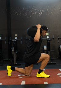 Uomo con maglietta nera e pantaloncini esegue un affondo, tenendo una sacca di sabbia nera sulle spalle, indossando scarpe da allenamento giallo brillante su un tappetino rosso.