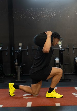 Hombre con camiseta y pantalones cortos negros realiza una zancada, sosteniendo una bolsa de arena negra sobre sus hombros, con zapatillas de entrenamiento amarillo brillante sobre una alfombra roja.