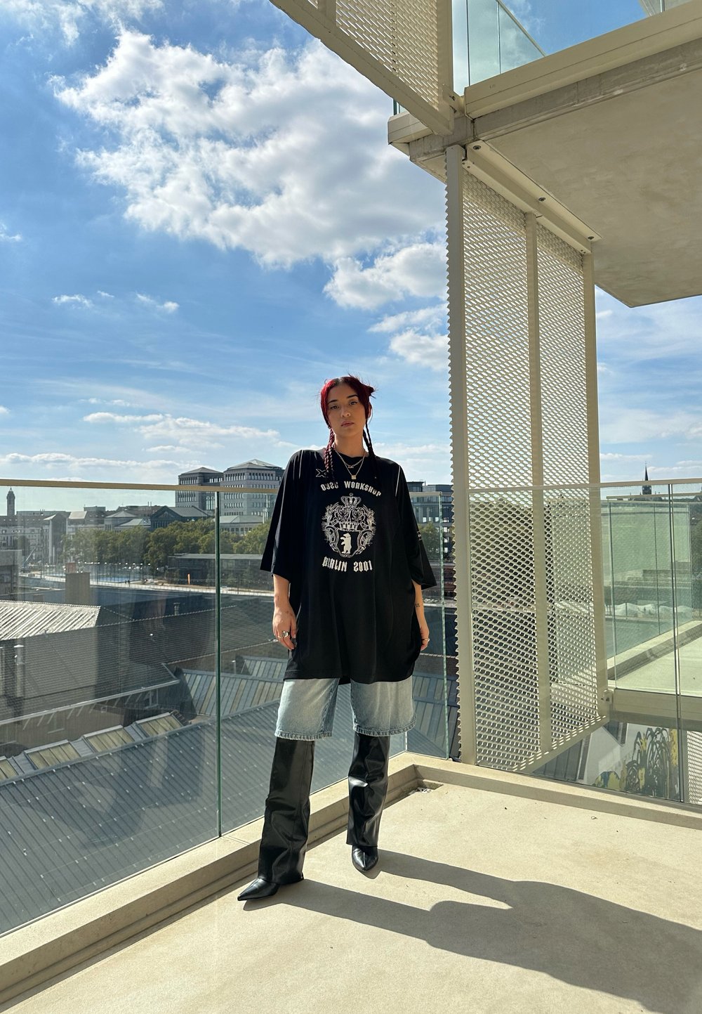 T-shirt oversized preta com uma estampa gráfica, combinada com calções de ganga azul claros e botas altas pretas brilhantes, contra um céu claro.