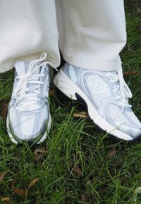 Białe i szare buty sportowe z siateczki i materiałów syntetycznych, z wyściółką w okolicy kostki, sznurówkami oraz teksturowaną podeszwą, umieszczone na trawie.