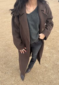 Γυναίκα που φοράει μακρύ καφέ παλτό, σκούρο γκρι πουλόβερ, μαύρα τζιν, καφέ μπότες και κρατά μια καφέ τσάντα σε μια επιφάνεια από χαλίκι.