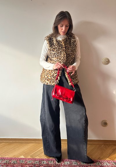 Chaleco de piel sintética de leopardo, camisa blanca de manga larga, pantalones anchos negros y un bolso de cuero rojo con un detalle de lazo y un asa voluminosa.