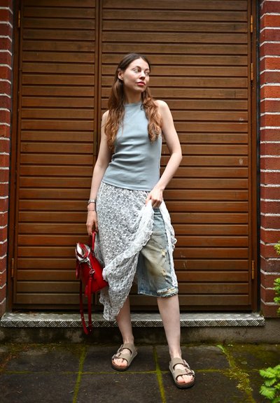 Top gris sin mangas, pantalones cortos de mezclilla desgastados, encaje blanco, bolso rojo y sandalias color tan, contra un fondo de tablones de madera.