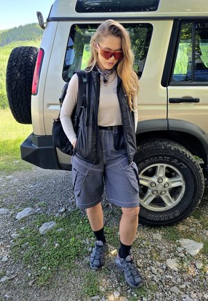 Una persona che indossa un gilet trapuntato nero, una maglia a maniche lunghe chiara, pantaloni cargo grigi e sandali da trekking neri si trova accanto a un veicolo.