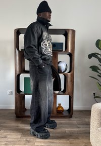 Homme portant un bonnet noir, une veste graphique, un jean large délavé et des bottes à semelles épaisses, debout à l'intérieur près d'une étagère en bois avec des objets décoratifs.