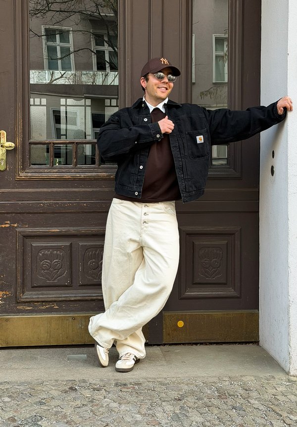 Άντρας με μαύρο μπουφάν, μπεζ παντελόνι, καφέ καπέλο και γυαλιά ηλίου ακουμπά σε καφέ πόρτα, χαμογελώντας με σταυρωμένα πόδια πάνω σε πέτρινο πλακόστρωτο.