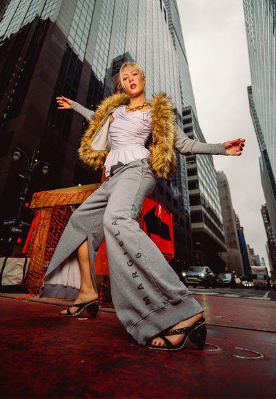 Mujer rubia con pantalones anchos grises y chaqueta con ribete de piel posa en una calle de la ciudad con altos edificios de cristal y tráfico al fondo.
