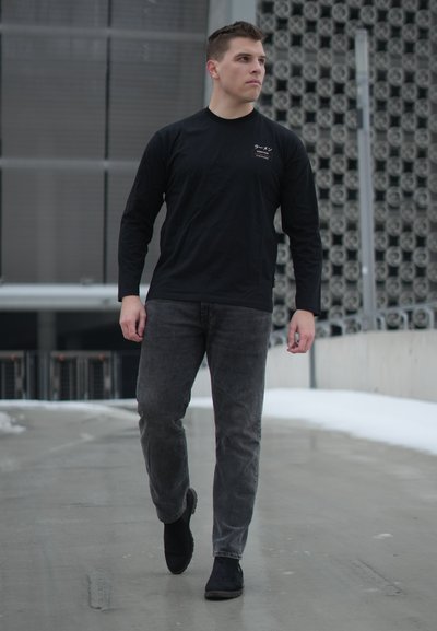 Joven caminando por la acera urbana, vistiendo una camiseta negra de manga larga con texto en japonés y jeans oscuros, mirando hacia la derecha.