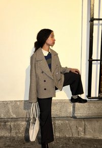 Manteau à double boutonnage en tartan marron et crème, superposé à une chemise rayée bleu marine, associé à un pantalon évasé noir et des chaussures brillantes, portant un sac blanc.