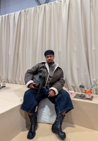 Chaqueta de cuero marrón con forro de piel, suéter gris, vaqueros azules y botas negras hasta el tobillo. Los accesorios incluyen un bolso y artículos decorativos.