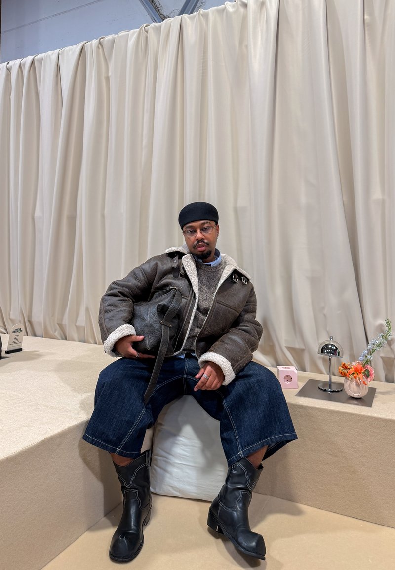 Bruine leren jas met schapenvachtvoering, grijze trui, blauwe jeans en zwarte enkellaarsjes. Accessoires omvatten een tas en decoratieve items.