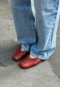 Κόκκινα δερμάτινα πέδιλα χωρίς λουράκι με στρογγυλή μύτη και σχέδιο με κοψίματα, συνδυασμένα με ανοιχτό μπλε τζιν παντελόνι, τοποθετημένα πάνω σε μια γκρι πέτρινη επιφάνεια.