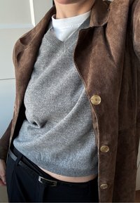 Long manteau en suède marron avec des boutons dorés, porté sur un pull gris clair. Col roulé blanc visible en dessous. Pantalon noir et ceinture.