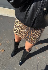 Crna kožna jakna, mini suknja s uzorkom leoparda i teksturiranom površinom, te visokotepene sjajne crne čizme s oštrim vrhom na popločenoj površini.