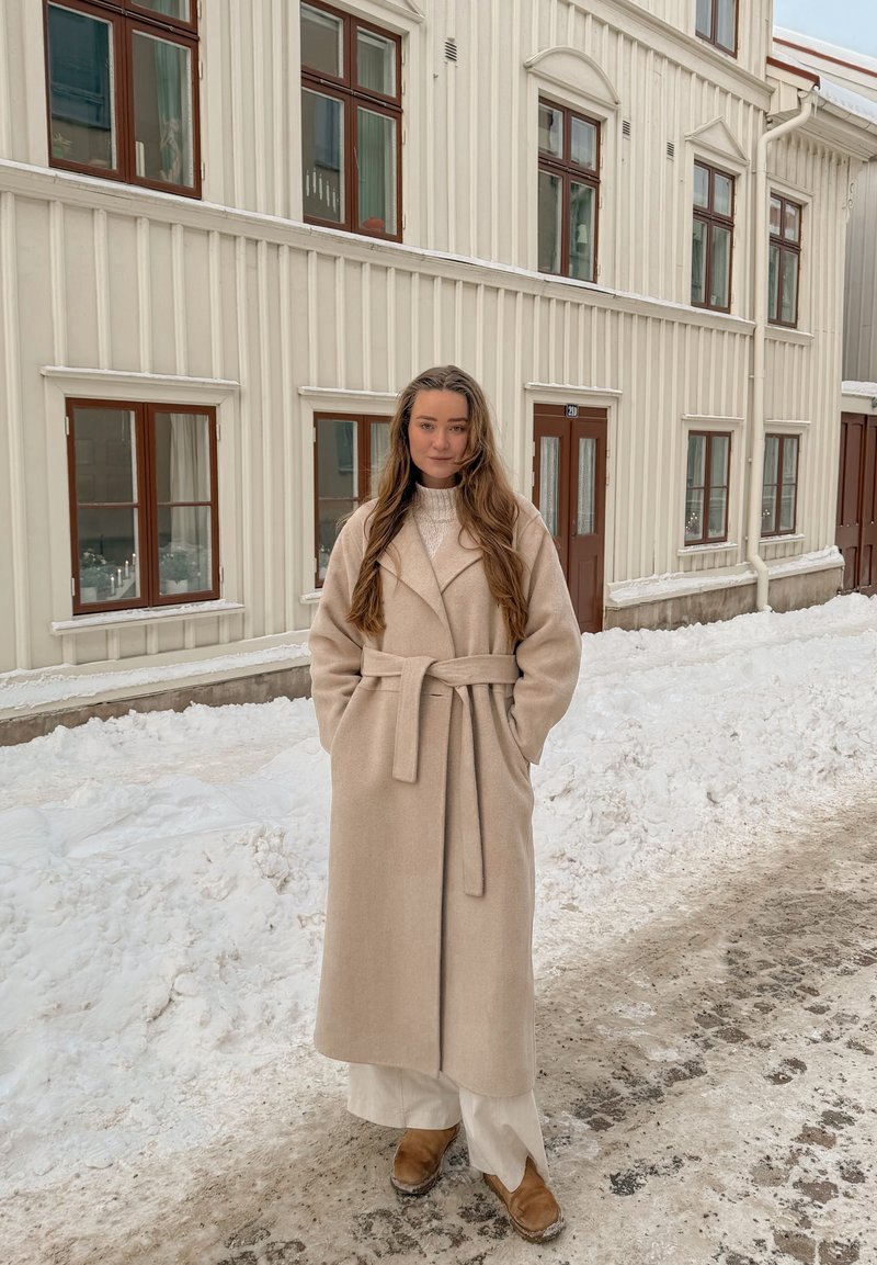 Femme en long manteau beige et bottes debout dans une rue enneigée devant un bâtiment en bois crème avec des fenêtres et des portes marron.