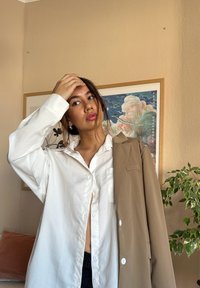 Λευκό oversized πουκάμισο με κουμπάκι και μία τσέπη, συνδυασμένο με καμηλό σακάκι κρατημένο στον έναν ώμο. Το φόντο περιλαμβάνει ένα πράσινο φυτό.