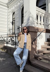 Giacca di tela beige con colletto marrone, T-shirt bianca, jeans a gamba larga azzurri e sneakers grigie; tiene in mano una tazza di caffè, in piedi vicino alle scale.