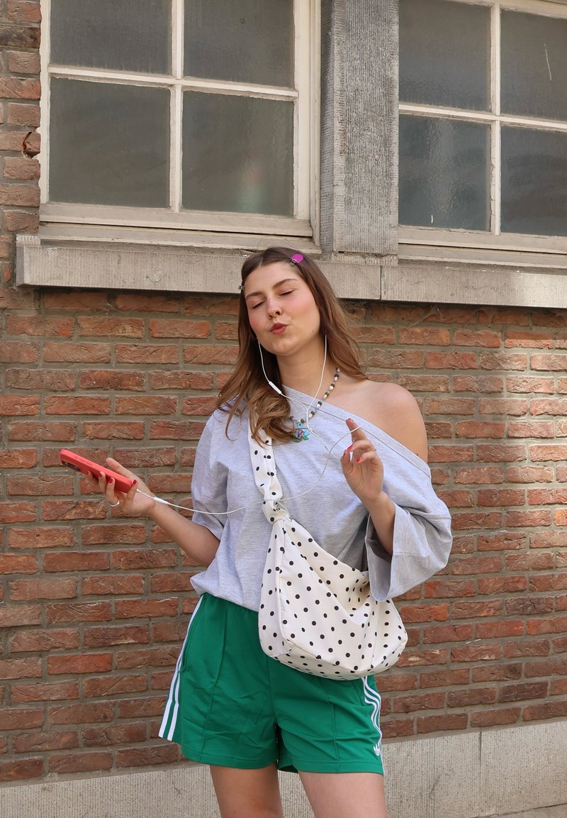 Young woman in green shorts and off-shoulder top listens to music with earphones, holding a phone and standing against a brick wall.