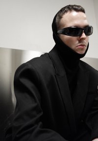 Blazer noir surdimensionné porté sur un col roulé noir, surmonté d'une capuche tricotée noire. La silhouette porte des lunettes de soleil foncées, avec un arrière-plan neutre.
