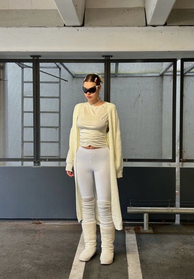 Leggings blancos ajustados combinados con una blusa de crema clara con fruncidos y un cardigan largo de punto. Botas blancas gruesas completan el atuendo. Se llevan gafas de sol negras.
