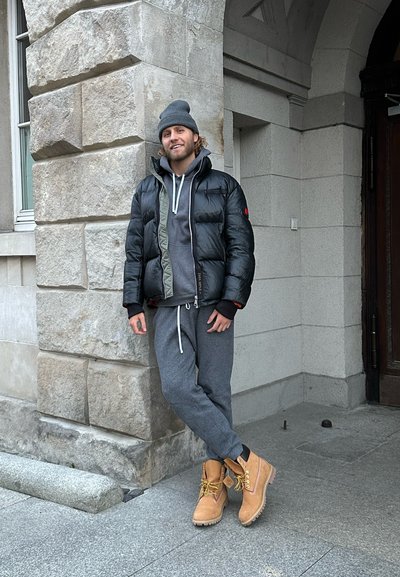 Chaqueta de plumas negra con un interior verde, sudadera gris debajo, pantalones de chándal grises y botas de piel color beige con cordones. Fondo de pared de piedra.
