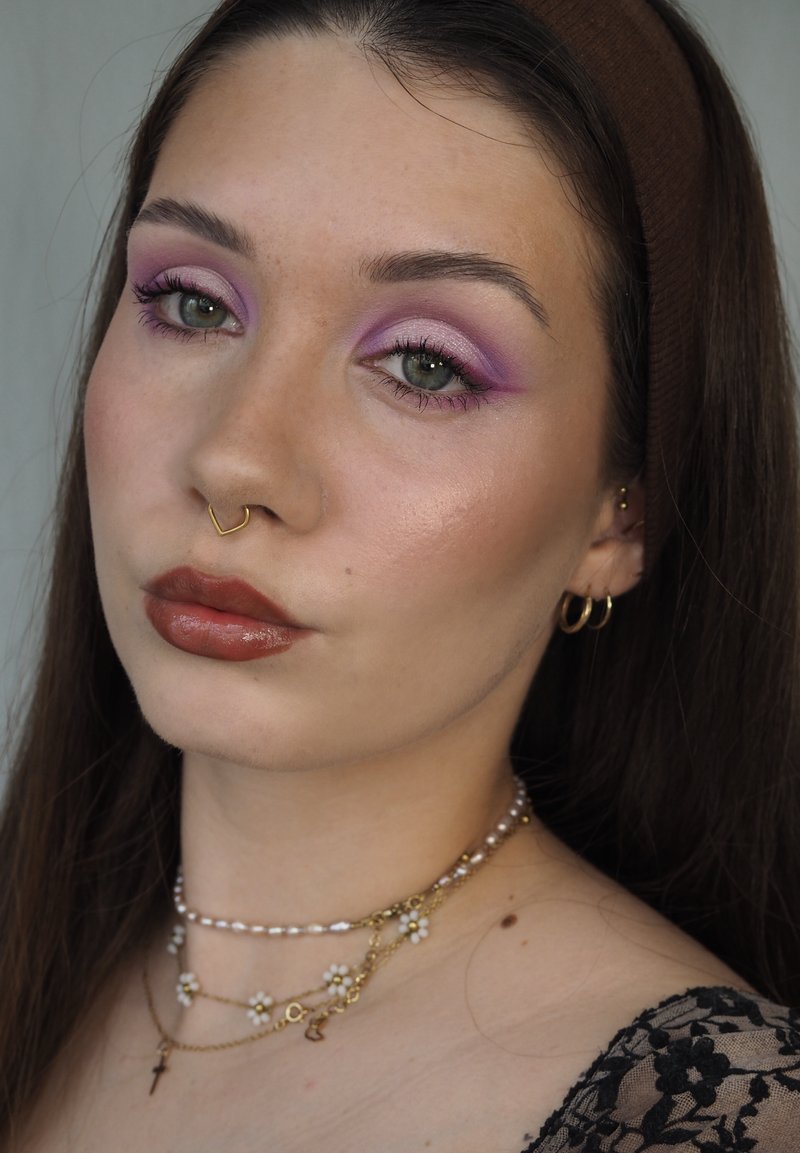 Jeune femme aux longs cheveux bruns, fard à paupières violet, rouge à lèvres marron, anneau de septum, boucles d'oreilles anneaux dorés, colliers superposés de perles et de fleurs dorées, haut en dentelle noire.