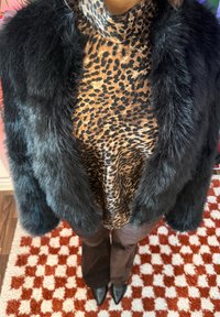 Personne portant un manteau en fausse fourrure noire, un col roulé léopard, un pantalon marron et des chaussures pointues noires, debout sur un tapis à carreaux rouges et blancs.