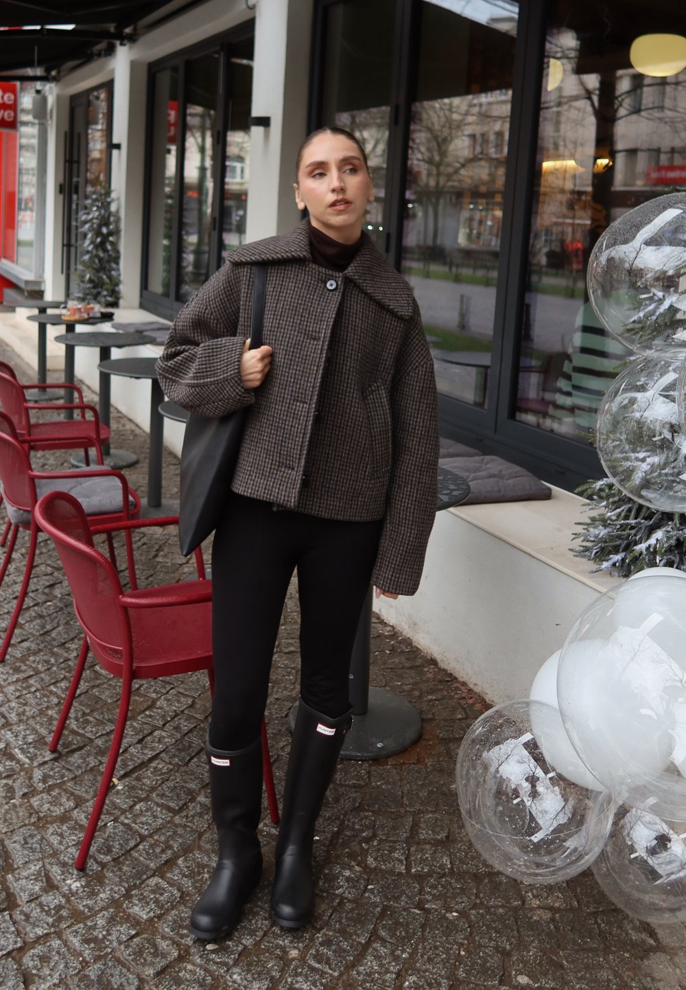 Femme en veste à carreaux, leggings noirs et bottes, debout près d'une terrasse de café en plein air avec des chaises rouges et des ballons transparents décoratifs.