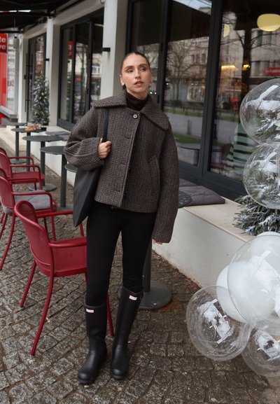 Mujer con chaqueta de cuadros, leggings negros y botas, de pie junto a la zona de asientos al aire libre de una cafetería con sillas rojas y globos transparentes decorativos.