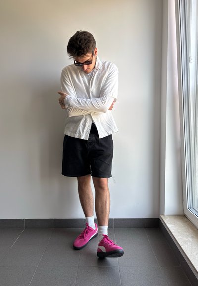 Camisa blanca de manga corta, pantalones cortos negros y zapatillas rosas con detalles blancos. De pie en un entorno minimalista interior con baldosas grises.