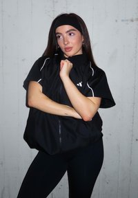 Fekete, rövid ujjú széldzseki fehér díszítésekkel és magas gallérral; a modell keresztbe tett karokkal pózol. A fekete leggings egészíti ki az öltözéket.