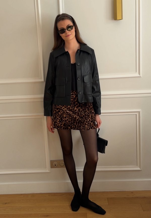 Femme portant une veste en cuir noire, une mini-jupe imprimé léopard, des collants noirs, des chaussures noires, des lunettes de soleil et tenant un petit sac à main noir à l'intérieur.