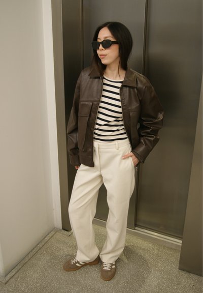 Mujer con gafas de sol negras, chaqueta de cuero marrón, camisa de rayas, pantalones blancos y zapatillas beige, de pie cerca de una puerta metálica de ascensor.