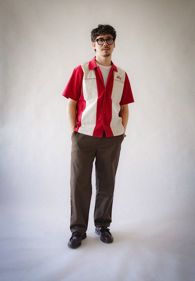 Camisa de manga corta roja y blanca con bolsillos frontales, combinada con pantalones marrones y zapatos negros. La camisa presenta una estética de diseño vintage.