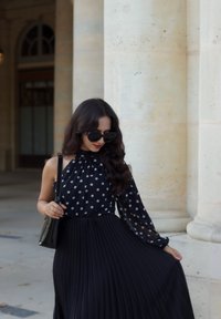 Blusa nera a pois con una manica e gonna nera plissettata. Capelli lunghi e mossi e occhiali da sole neri. Tiene una piccola borsa nera.