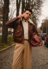 Veste en cuir marron avec col en shearling, pull crème en maille torsadée, pantalon beige à jambes larges et sac en cuir rouge, posés sur un chemin pavé.