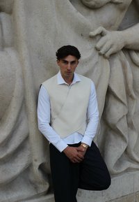 Kremo barve, jopič v ovitku čez belo srajco, v kombinaciji s temno prugastimi hlačami. Lahek videz, stoji ob ozadju teksturiranega kamenja.