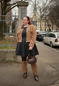 Femme aux cheveux roux debout dans la rue, portant une robe noire, une veste courte couleur bronze, des guêtres en fourrure marron, et tenant un sac à main marron.