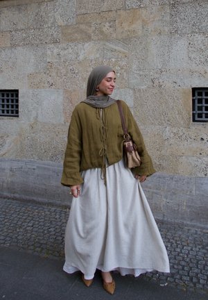 Chaqueta corta de lino suelta en verde oliva con lazo al frente, combinada con una falda fluida de un beige claro. Zapatillas de tacón bloque trenzadas en color beige y bolso.