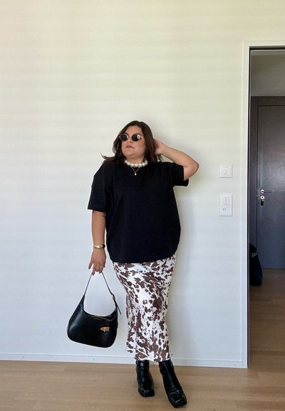 Camiseta negra de gran tamaño, falda midi de manchas blancas y marrones, bolso negro y botas de tacón negras. Los accesorios incluyen un collar de perlas y gafas de sol.