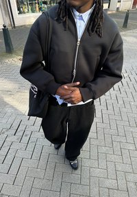 Schwarze Bomberjacke mit Reißverschluss und zwei Seitentaschen, gestreiftes Button-Down-Hemd darunter, lockere schwarze Hose und schwarze Schuhe.
