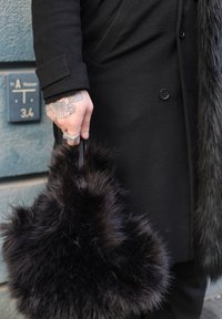 Sac à main en fausse fourrure noire de forme arrondie, avec une texture douce et des poignées en cuir, tenu par une main avec un tatouage et une bague argentée.