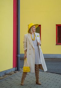 Μπεζ παλτό, γκρι φόρεμα με υφή, κίτρινο καπέλο δήλωσης και κολιέ, καθώς και κίτρινη τσάντα με αλυσίδα. Καφέ μποτίνια μέχρι το μηρό.