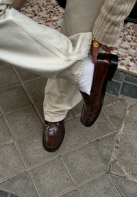 Brune læder loafers med en glat finish, detaljeret syning og en sort gummisål, båret med hvide sokker og cremefarvede bukser.