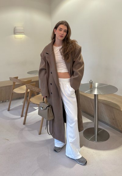 Abrigo marrón de gran tamaño, top blanco corto, pantalones blancos, zapatillas grises y bolso gris. Interior moderno con muebles de madera y una mesa de metal.