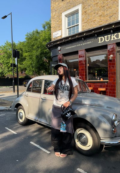 Coche gris vintage con faros redondos aparcado al lado de un pub de ladrillo. Persona con camiseta gráfica, pantalones grises y bolso negro está cerca.