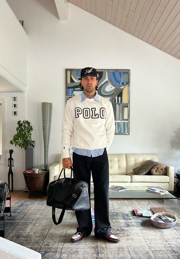 Man i casual outfit med POLO-tröja, svart keps, som håller en svart läderväska, står i ett modernt vardagsrum med konst och soffa bakom sig.
