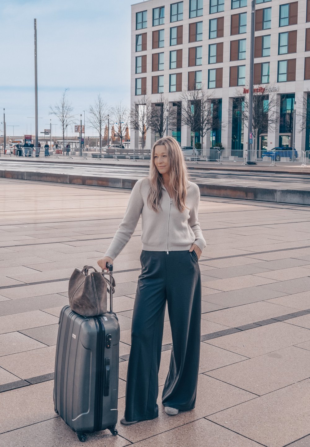 Valise à roulettes noire à surface rainurée à côté d'une personne portant un pull zippé gris clair et un pantalon large foncé, tenant un sac à main marron.