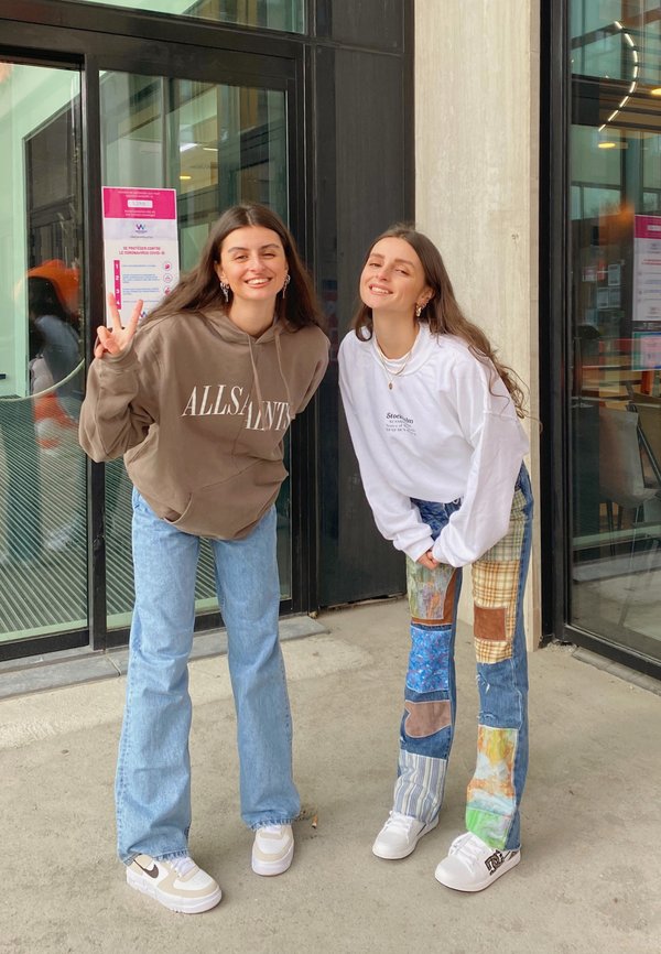 Dos personas están de pie frente a unas puertas de cristal. Una lleva una sudadera marrón y jeans azules; la otra lleva una camisa blanca de manga larga y pantalones de retazos.