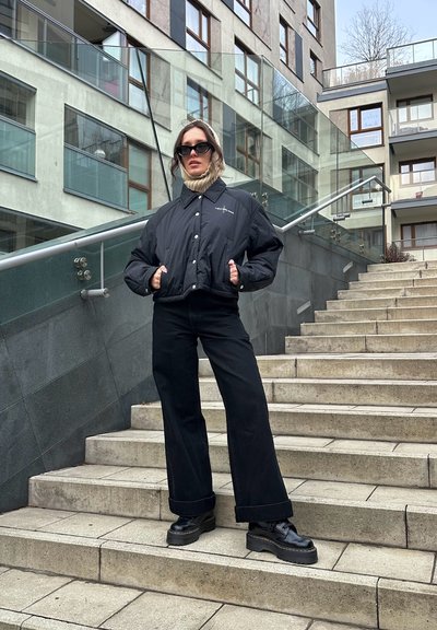 Chaqueta de plumas negra con diseño oversize, cuello alto y botones frontales. Combinada con pantalones negros de cintura alta y zapatos negros de plataforma.